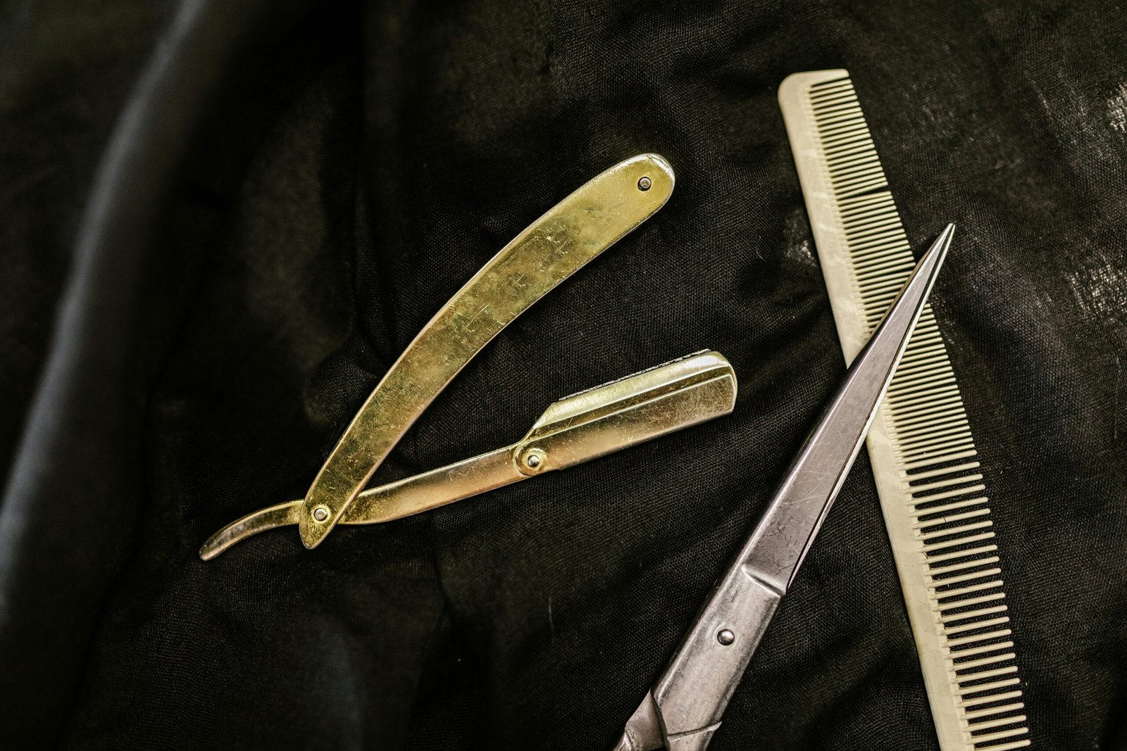 Close-up of silver shears, comb, and straight razor on a black cloth background, highlighting barber tools.